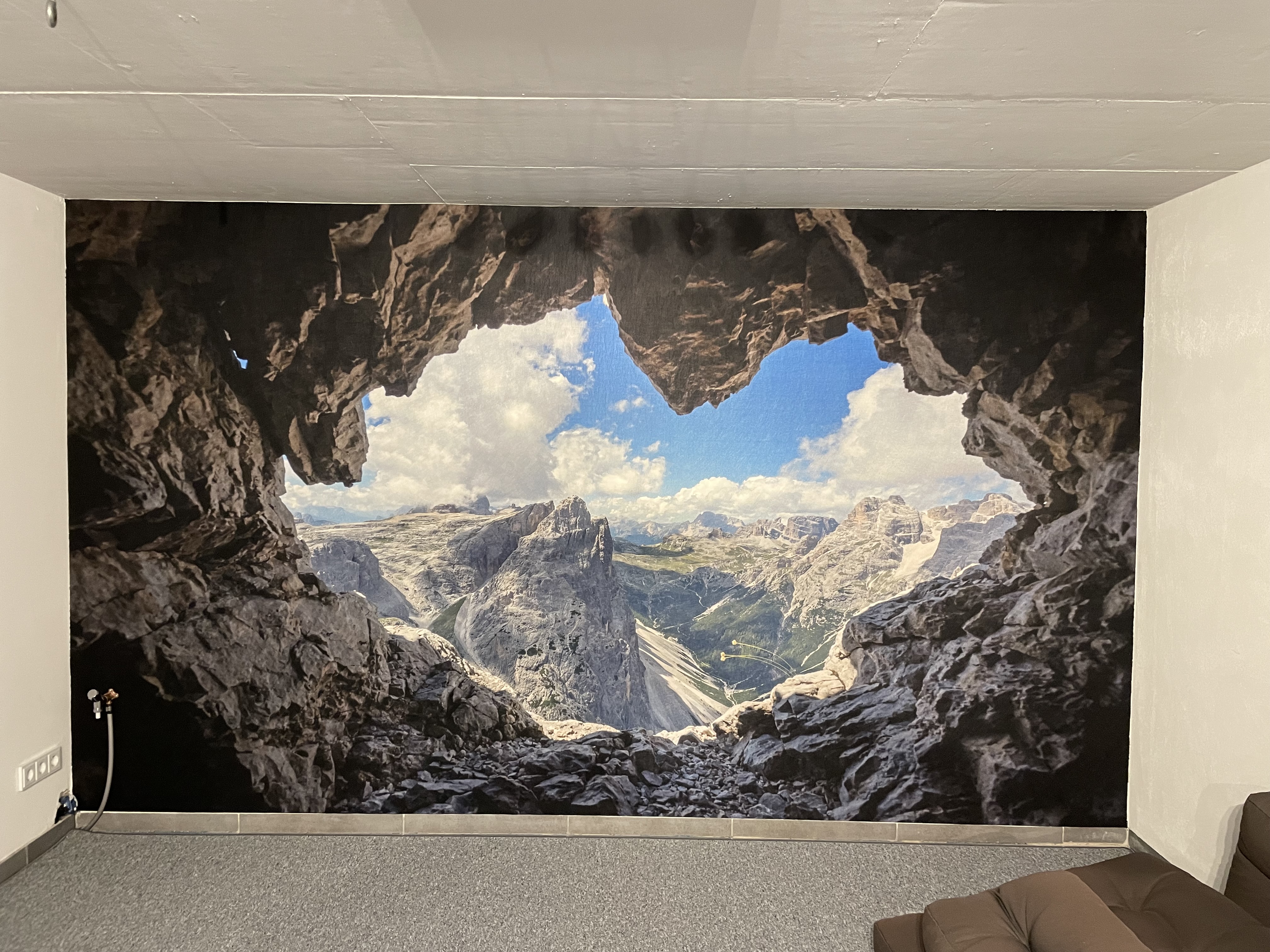 Großformatige Fototapete an der Wand mit Blick aus einer Felsenhöhle auf beeindruckende Alpenberge, blauen Himmel und Wolken.