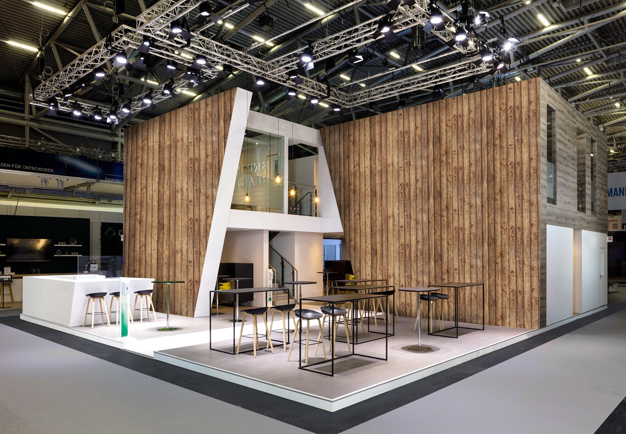 Großer Messestand mit Holzoptik-Wänden, weißem Empfangsbereich, Tischen und Barhockern als Beispiel für professionellen Messebau